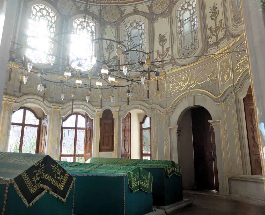 Şah Sultan Türbesi'nin içi; yeşil örtülü sandukalar, kalem işi duvarlar, yaldızlı yazı kuşağı ve renkli camlı pencereler.