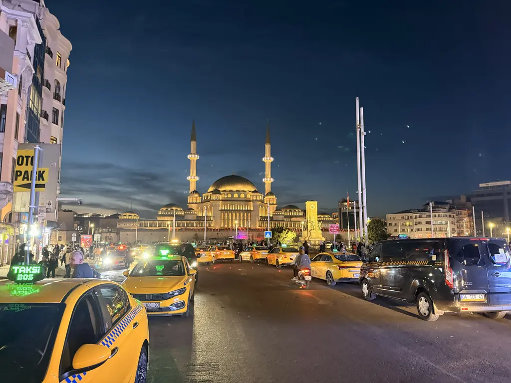 Gece Taksim Meydanı'na çıkan caddede sarı taksiler; ileride ışıklandırılmış Taksim Camii ve iki minaresi.