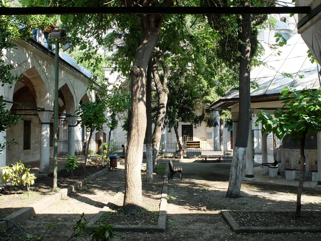 Nişancı Mehmed Paşa Camii'nin avlusu; kemerli revaklar, gölge veren ağaçlar, taş döşeli yol ve sağda şadırvanın saçağı.