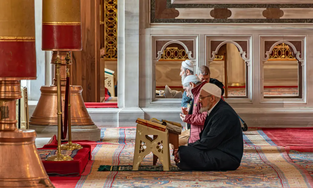 Fatih Camii'nde halının üzerinde oturan yaşlı bir adam ahşap rahledeki Kur'an'ı okuyor; solda kırmızı abajurlu pirinç şamdan, arkada beyaz takkeli iki adam ile bir çocuk.