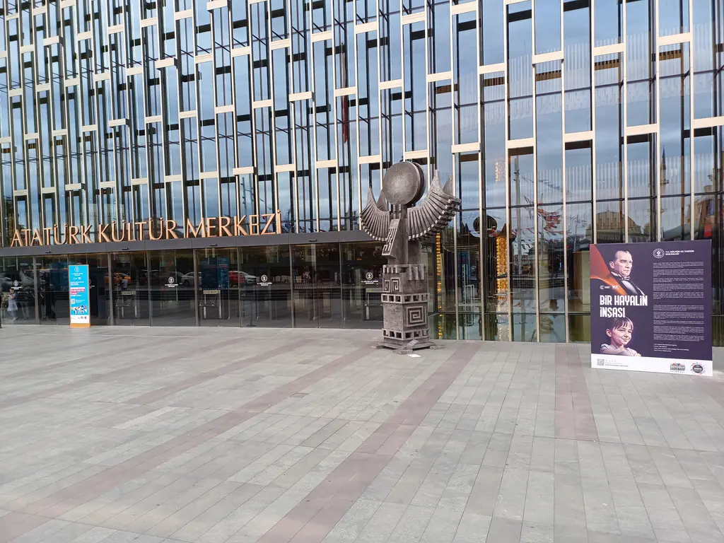 Atatürk Kültür Merkezi'nin girişi; önde taş meydan ve kahverengi metal heykel, arkada cam cephe, giriş kapıları ve afiş panoları.