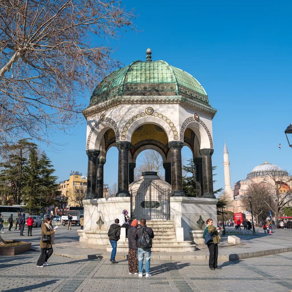 Alman Çeşmesi'nin merdivenli cephesi; basamaklar, kemerler arasından görünen mozaik, çevrede ziyaretçiler ve arkada Ayasofya'nın kubbesi.
