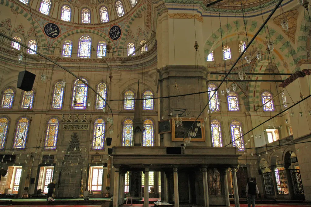 Fatih Camii'nde müezzin mahfili; kısa sütunlara oturan korkuluklu mermer platform, arkada kat kat vitraylı pencereler, boyalı kemer bantları ve siyah zeminli hat madalyonları.