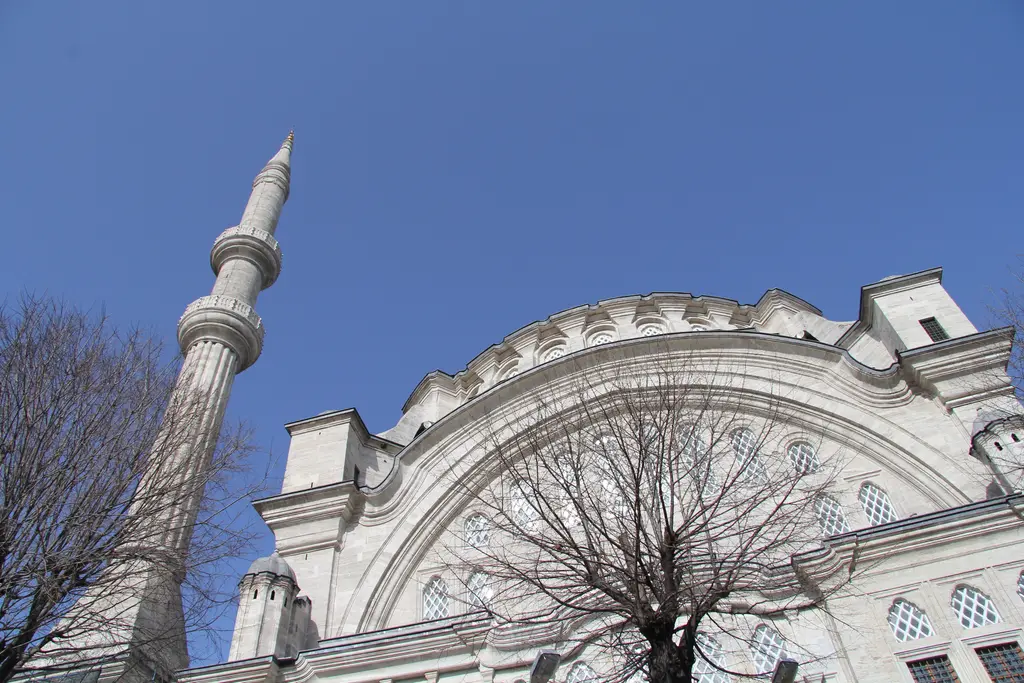 Nuruosmaniye Camii'nin cephesi alttan; duvarı baştan başa kaplayan dev kemer ile içindeki pencere ızgarası, solda minare gövdesi ve önde yapraksız ağaç dalları.