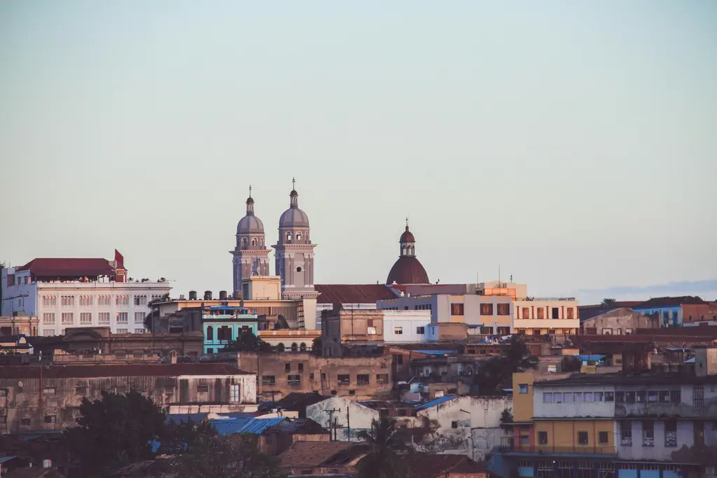 Santiago de Cuba'da şehir manzarası; öndeki alçak evlerin ardında yükselen çift kuleli katedral ile kubbeler, sıralanan renkli binalar ve puslu gökyüzü.