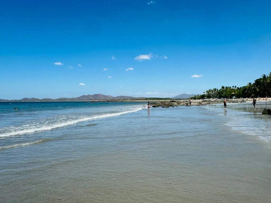 Tamarindo'da güneşli havada geniş kumsal; kıyıya doğru alçak dalgalarla gelen deniz, ıslak kumdaki yansımalar ve arkada denize uzanan ormanlık tepeler.
