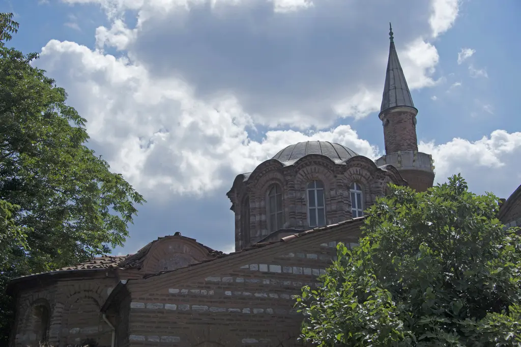 Vefa Kilise Camii'nin kubbesine ağaç tepeleri arasından yukarı bakış; yüksek kasnak, kemerli pencereler, dalgalı tuğla saçak, kurşun külah ve altta almaşık duvar.