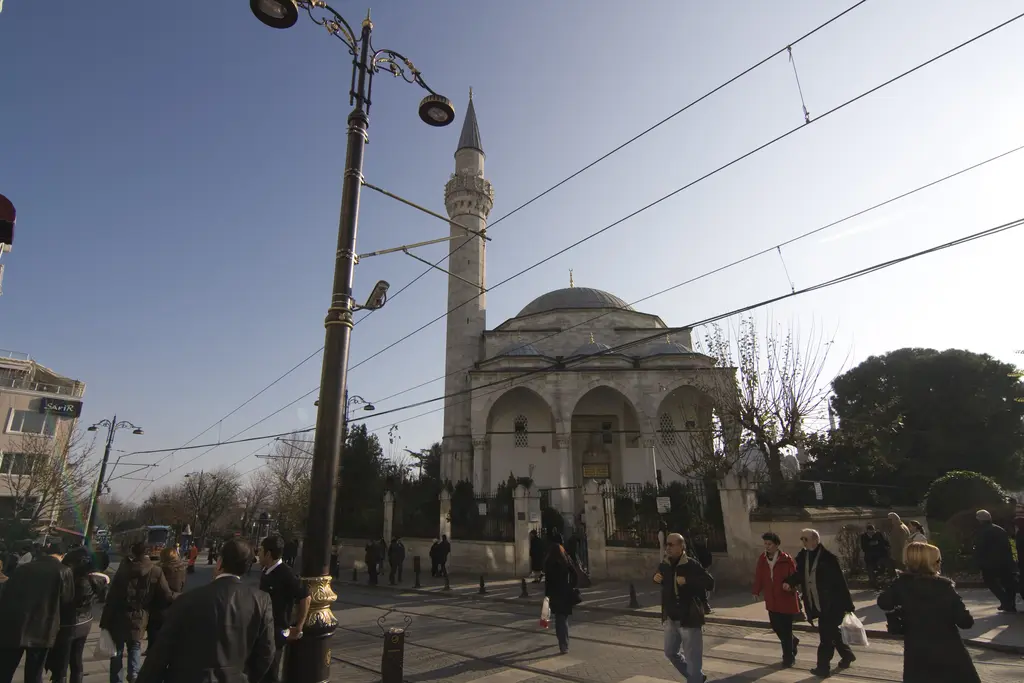 Divanyolu'nda Firuz Ağa Camii; önde yürüyen kalabalık, sokak lambası ve tramvay telleri, arkada kubbe ve minare.