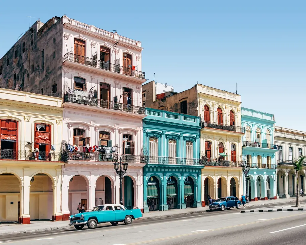 Havana'da pastel renkli sömürge binaları sırası; balkonlu cepheler önünde caddede ilerleyen turkuaz ve mavi klasik arabalar, berrak mavi gökyüzü.