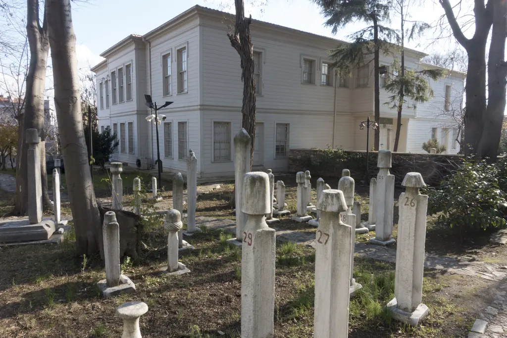 Hazirenin mezar taşları arasından mevlevihanenin ana binası; önde ağaçlar, arkada açık renkli iki katlı cephe.