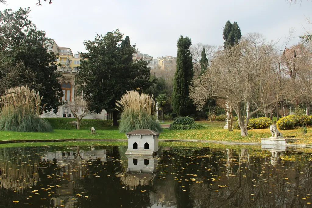 Ihlamur Kasrı'nda havuzun ortasında beyaz küçük bir kuş evi ve yansıması, kıyıda pampas otları, selviler ve mermer aslan heykeli.