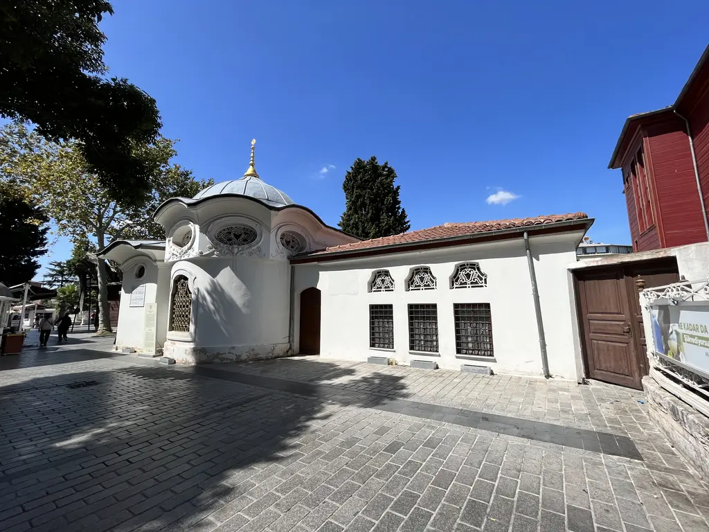 Koca Mustafa Paşa Külliyesi'nin avlusu; önde beyaz sıvalı, dalgalı saçaklı ve kurşun kubbeli türbe, sağ arkada kırmızıya boyalı iki katlı ahşap tekke binası, aralarda serviler.
