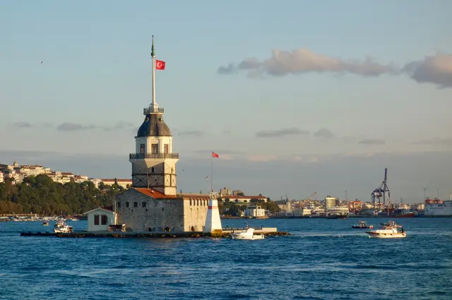Üsküdar açığında Kız Kulesi; küçük adacık üzerinde taş kule, tepesinde bayrak, çevresinde mavi deniz ve uzakta Avrupa yakası.