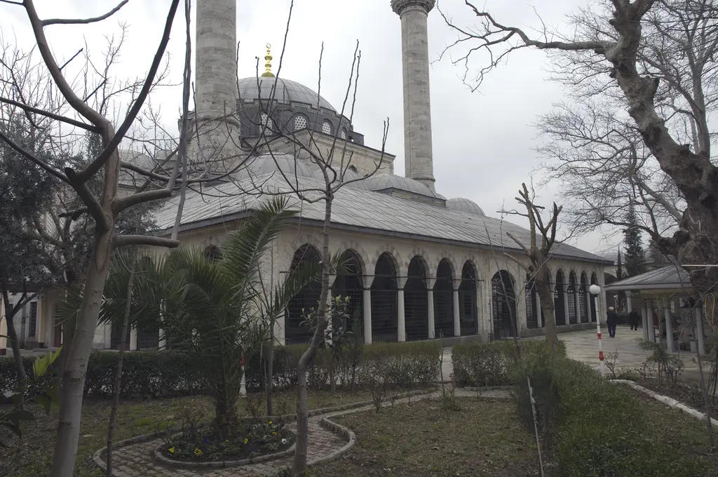 Atik Valide Camii; ağaçlı avlunun ardında kemerli revak, kurşun kubbe ve iki ince minare.