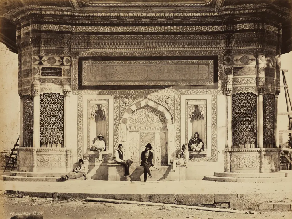 1867'de çekilmiş fotoğraf; çeşmenin bir cephesi, nişlerde ve sekilerde oturan adamlar, önünde toprak zemin.