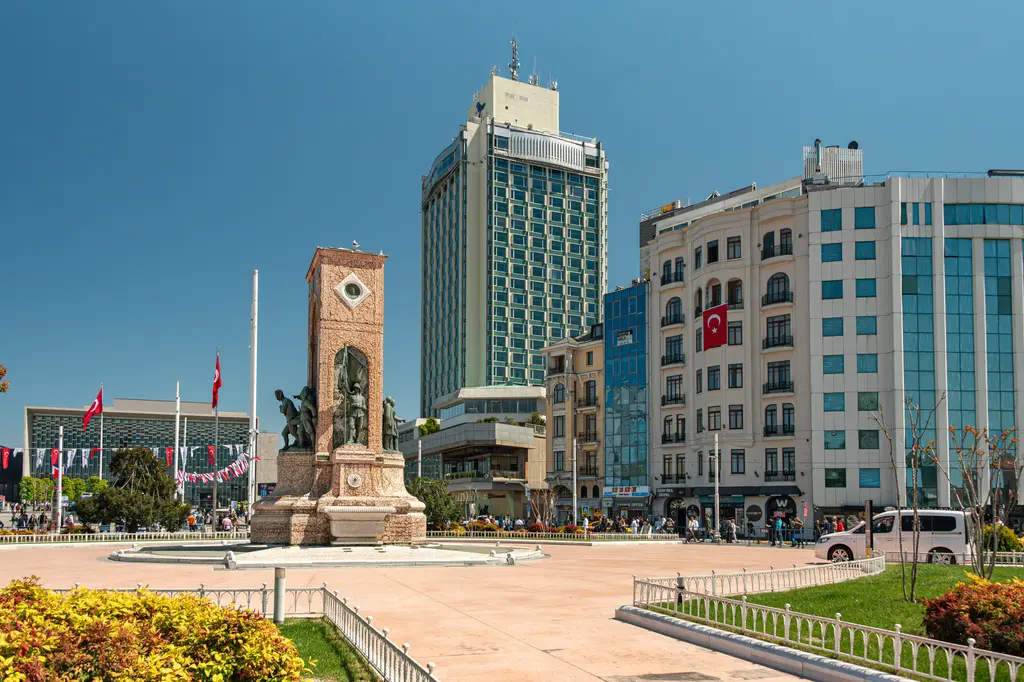 Cumhuriyet Anıtı ve çevresindeki çiçekli bahçe; arkada yüksek bir otel, sağda cephesi bayraklı çok katlı binalar.