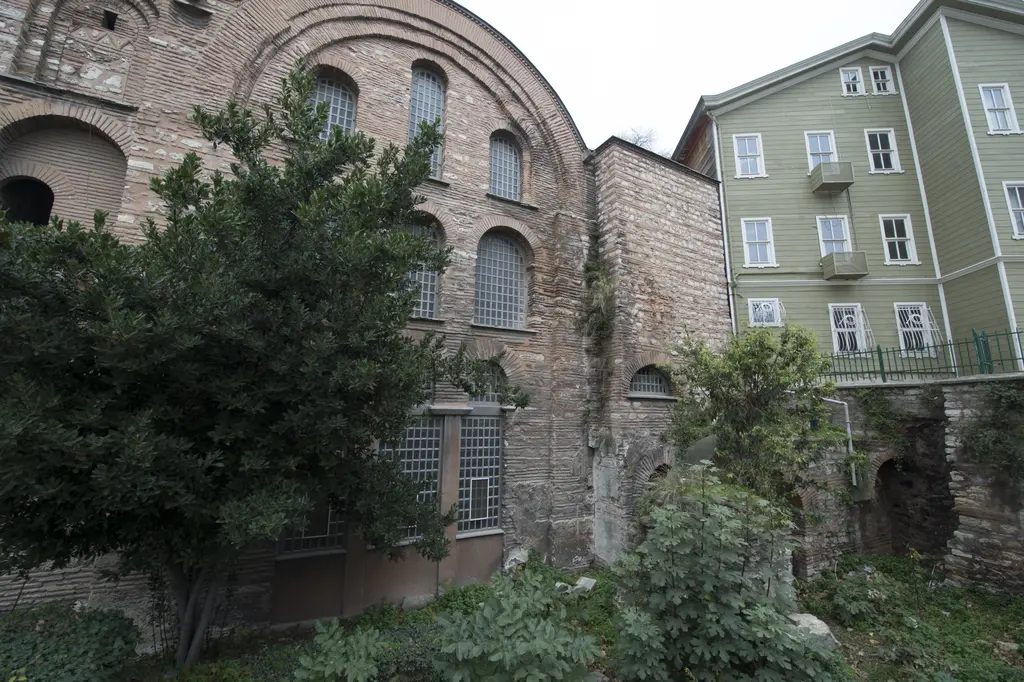 Kalenderhane Camii'nin güney yanı; yarım daire alınlıklı tuğla cephe, önde yoğun yeşil bir ağaç ve sağda yeşil boyalı ahşap ev.