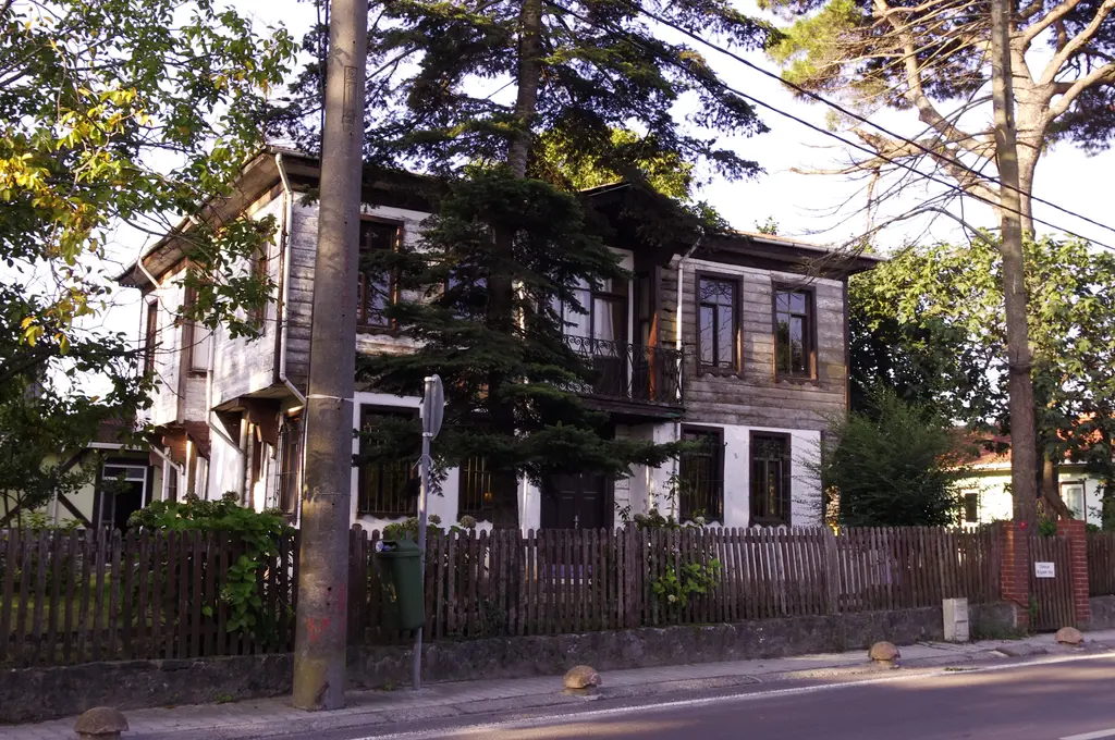Polonezköy'de iki katlı eski ahşap ev; çıkmalı üst kat, beyaz pencereler, önünde ahşap çit, çam ağaçları ve asfalt yol.