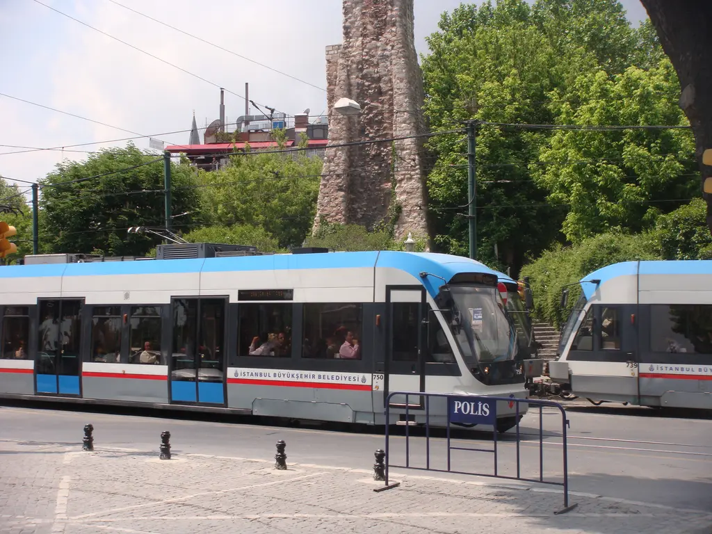Milyon Taşı'nın önünden geçen T1 tramvayı; arkada su terazisinin kulesi, yeşil ağaçlar ve önde bariyerli taş kaldırım.