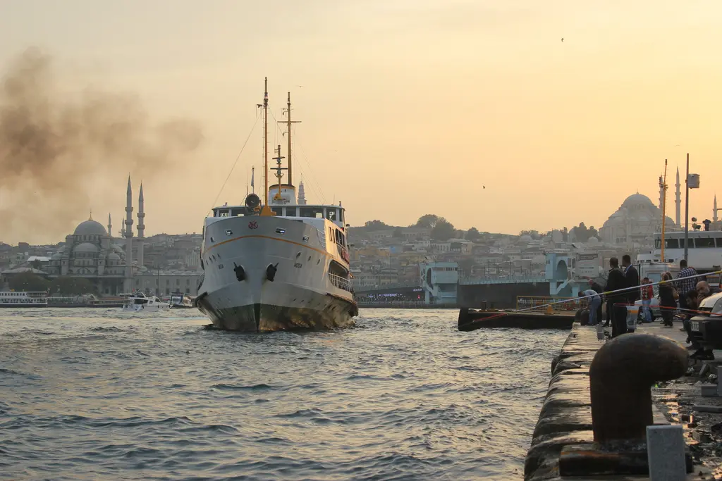 Akşamüstü Karaköy iskelesine yaklaşan vapur; dumanlı gökyüzü, rıhtımdaki babalar ve uzakta cami siluetleri.