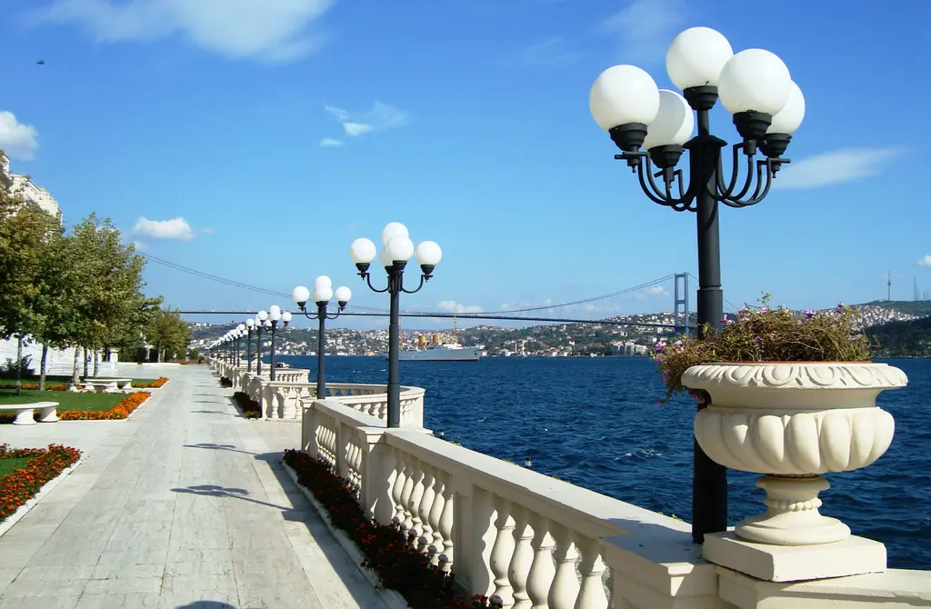 Çırağan Sarayı'nın sahil terası; taş döşeli yol, mermer korkuluk, beş kollu fenerler, arkada Boğaz ve köprü.