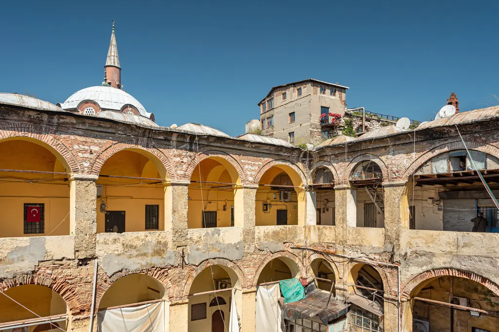 Büyük Yeni Han'ın avlu revakları; sarı boyalı duvarlar, tuğla örgülü kemerler, üstte komşu yapının kubbesi ve minaresi.