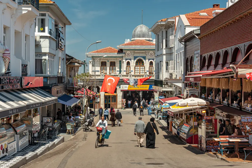 Büyükada çarşısı; kafeler ve dükkânlarla çevrili yaya caddesi, yürüyen insanlar ve sonunda kubbeli iskele binası.