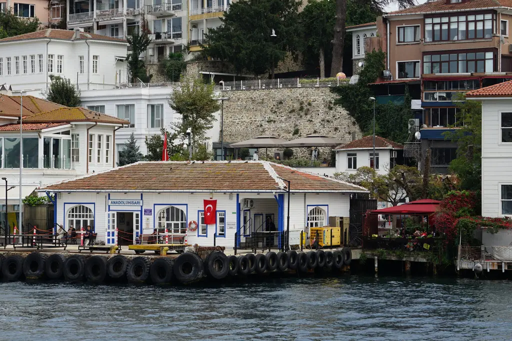 Anadoluhisarı iskelesi denizden; beyaz iskele binası, bayrak, arkada yalılar ve yamaçtaki sur.
