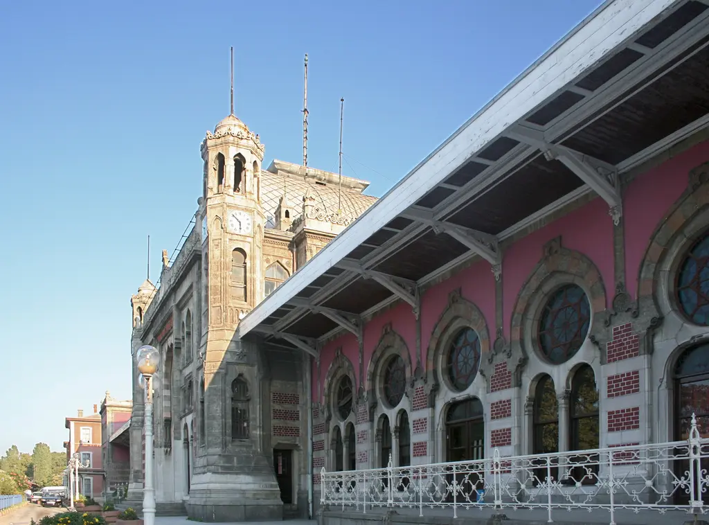 Sirkeci Garı'nın yan cephesi; solda taş saat kulesi, sağa doğru uzanan beyaz demir ve ahşap peron saçağı, altında pembe duvarlı arkad ve beyaz süslü korkuluk.