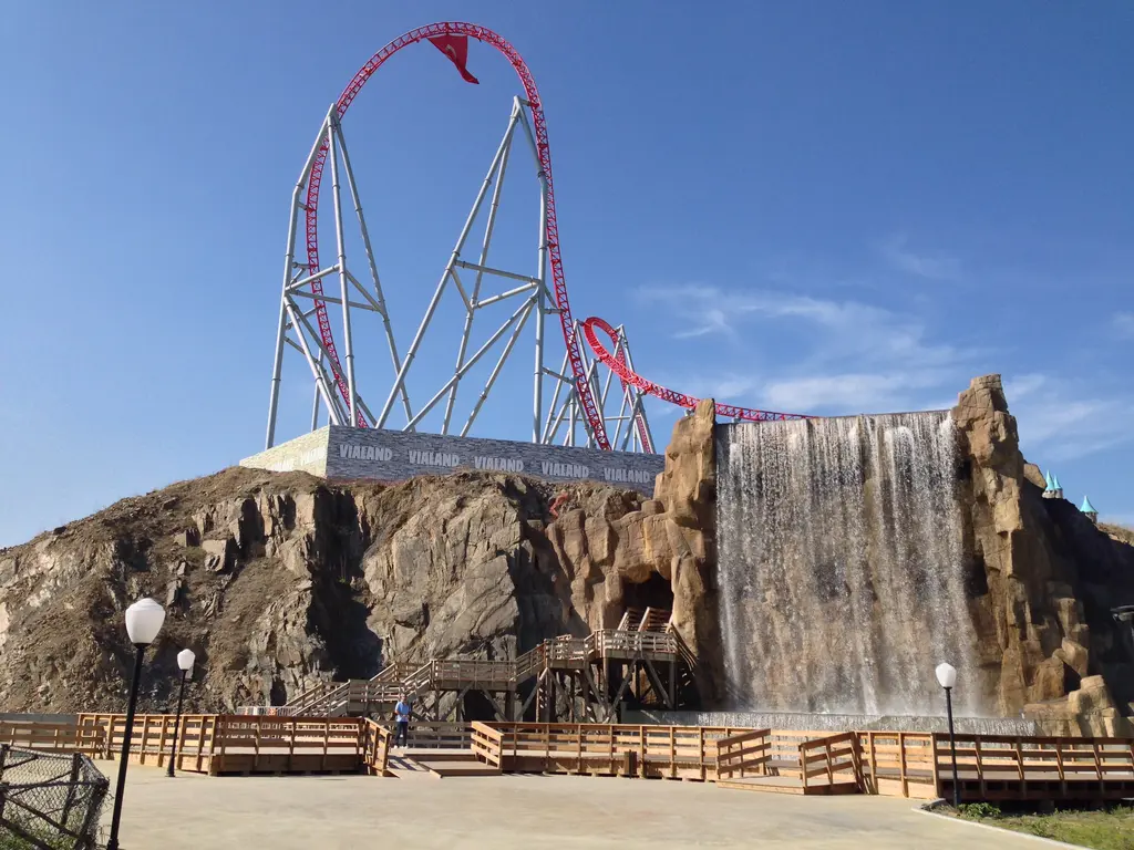 Vialand'de yapay kaya tepesi ve çağlayan; tepenin üstünde yükselen kırmızı roller coaster rayları, önde ahşap korkuluklu yol.