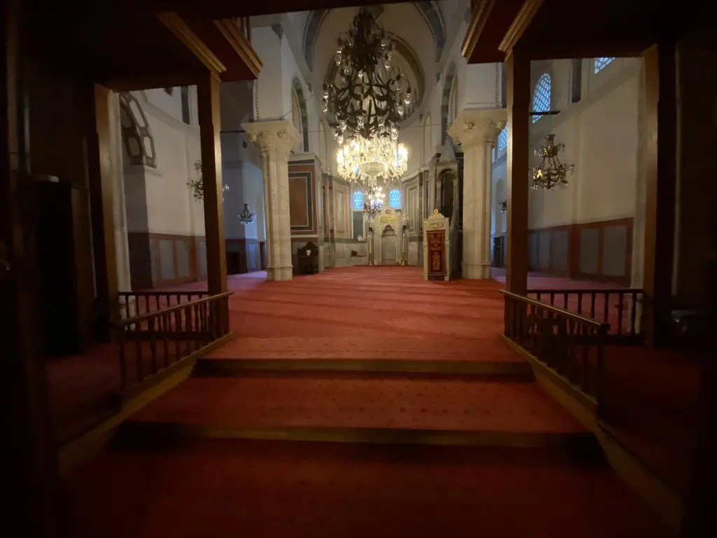 Zeyrek Camii'nde loş bir iç görünüm; ortada kırmızı halı kaplı geçit, iki yanda ahşap korkuluklar, tavanda kristal avize ve dipte aydınlık kemerli mekân.
