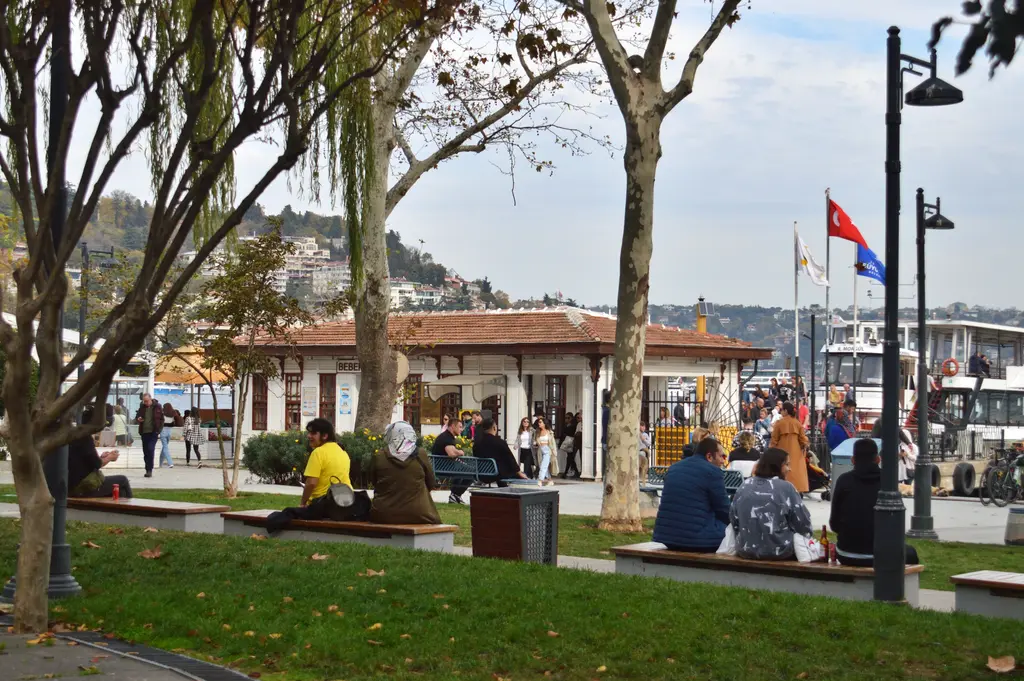 Bebek Parkı; çimenlik, ağaçlar, banklarda oturan insanlar, sokak lambaları ve arkada iskele binası.