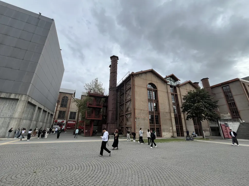 Santralistanbul'da yerleşke meydanı; solda gri sanat binası, ortada tuğla baca ve makine dairesi, taş zeminde öğrenciler.