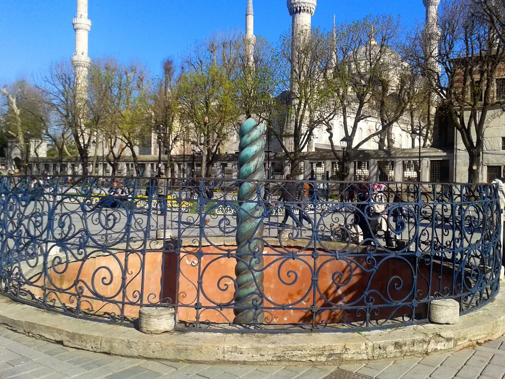 Yılanlı Sütun ve parmaklıklı çukuru; arkada Sultanahmet Camii'nin minareleri, yapraksız ağaçlar ve meydanda yürüyen insanlar.
