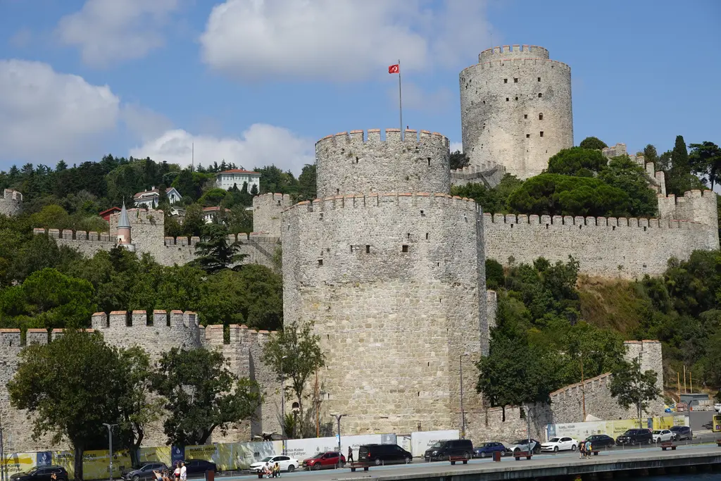 Rumeli Hisarı'nın kuleleri; büyük yuvarlak taş kuleler, mazgallı surlar, bayrak ve önünde sahil yolu.