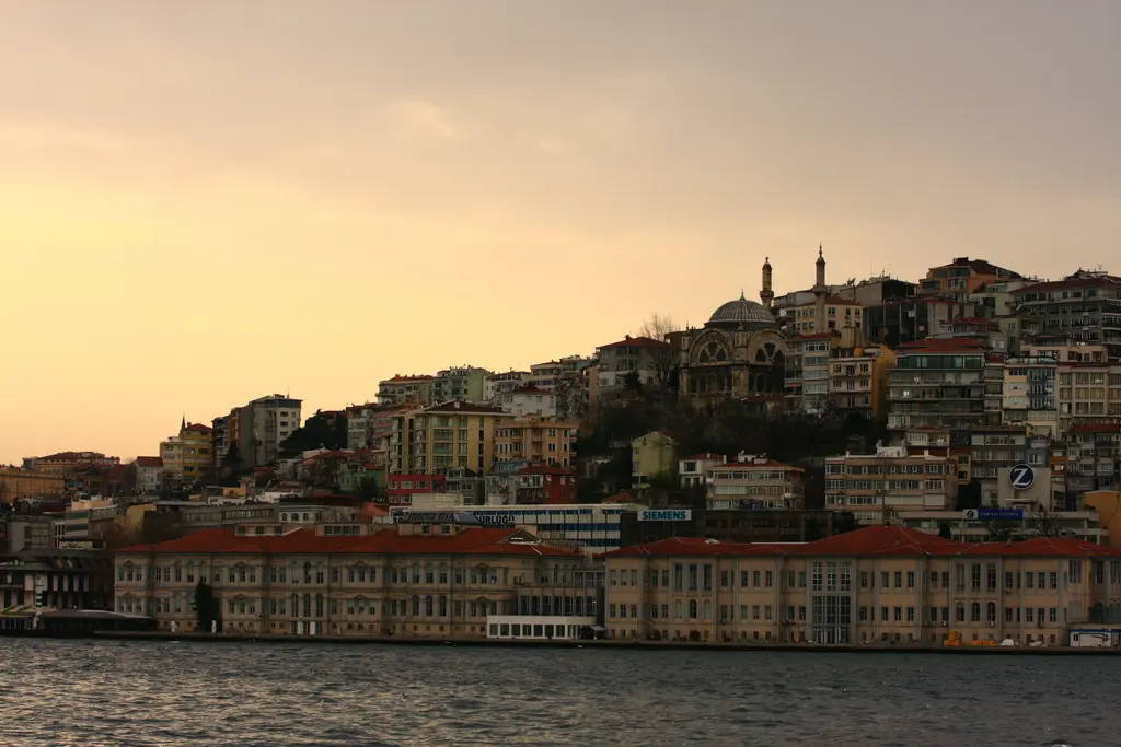 Gün batımında Cihangir yamacı; turuncu gökyüzü önünde cami siluetinin çevresinde evler ve kıyıda uzun tarihî binalar.