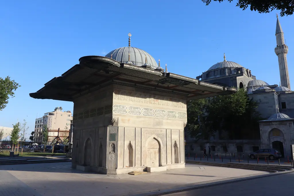 Tophane Çeşmesi'nin bir köşesi; geniş kurşun saçak, mermer gövdede nişler, arkada caminin kurşun kubbesi, minaresi ve mavi gökyüzü.