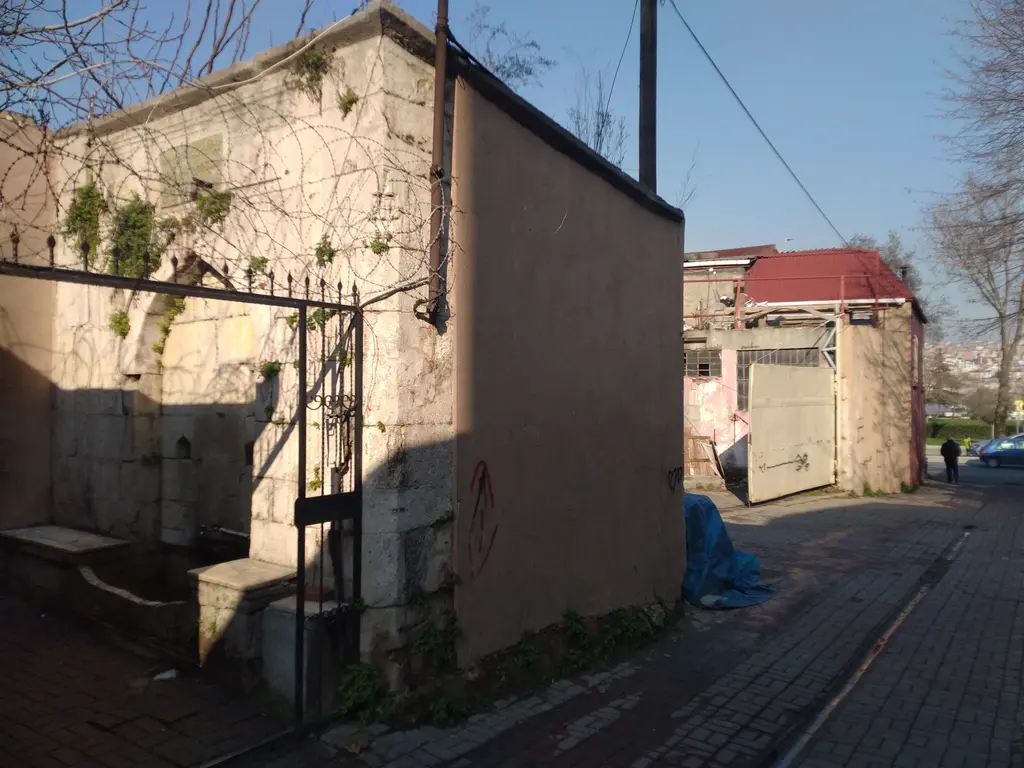 Ayvansaray'da Şatır Hasan Ağa Çeşmesi; demir kapılı küçük bir avlu içinde kesme taş çeşme gövdesi, önünde taş seki, yanında boyalı duvar ve dar Arnavut kaldırımı sokak.