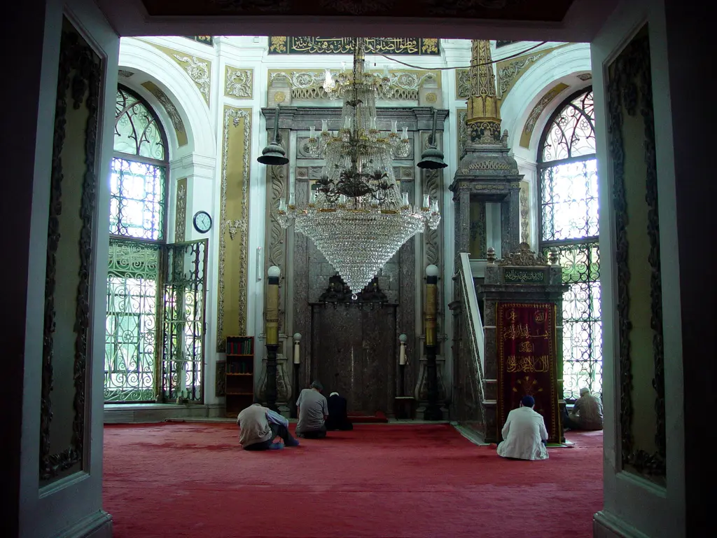 Zeyrek Camii'nde namaz vakti; kapıdan görünen harimde iki kişi secdede, kırmızı halı, yanda mihrap ile minber ve tavanda avizeler.
