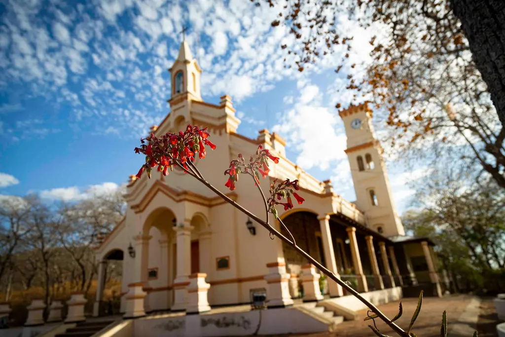 Paraguay'da sömürge üslubunda krem rengi bir kilise; çan kulesi ve saat kulesi, önünde kırmızı çiçekli bir dal, mavi-beyaz bulutlu gökyüzü.