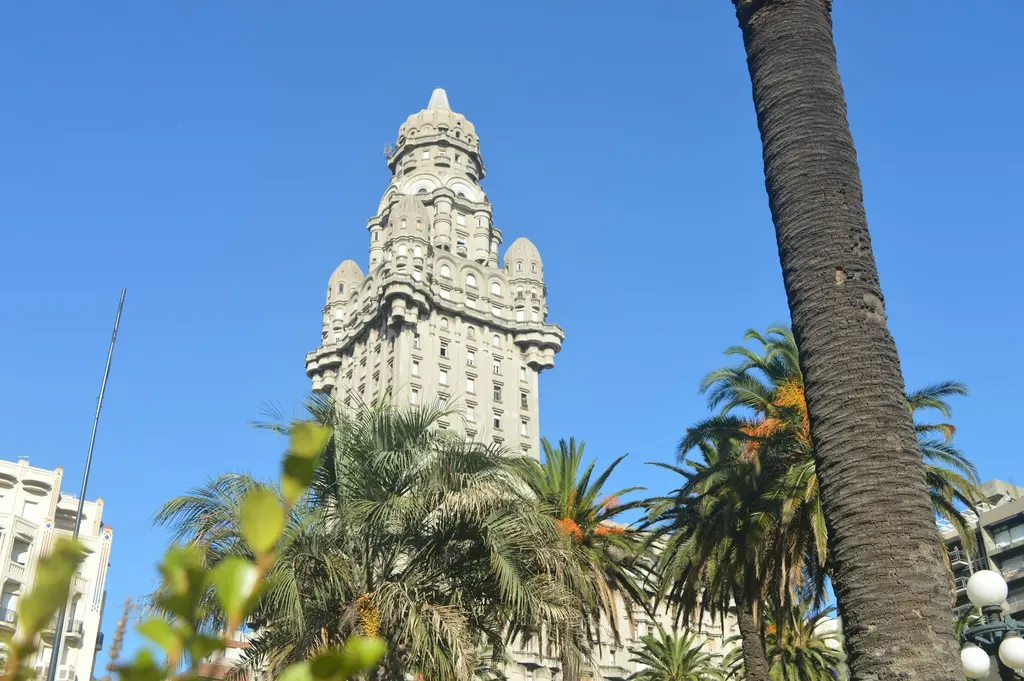 Uruguay'ın başkenti Montevideo'da Palacio Salvo'nun görkemli kuleli binası; önünde palmiyeler, berrak mavi gökyüzü altında aşağıdan çekilmiş görünüm.