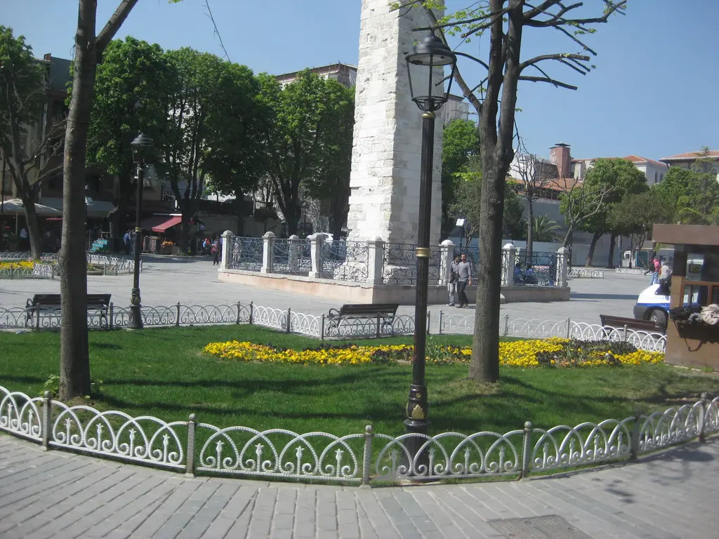 Örme Sütun'un alt kısmı; önde sarı çiçekli tarh ve çimen, beyaz parmaklık, ağaçlar, sokak lambası ve meydanda insanlar.
