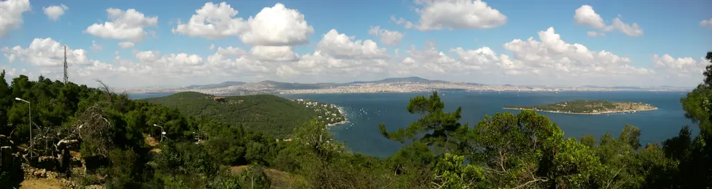 Yücetepe'den panorama; ormanlık yamaçlar, mavi Marmara, uzakta Sedef Adası ve bulutlu gökyüzü.