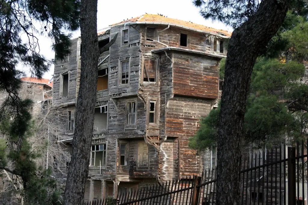 Büyükada Rum Yetimhanesi; çam dallarının ardında ahşap kanat, çatı saçağı ve önde demir parmaklık.