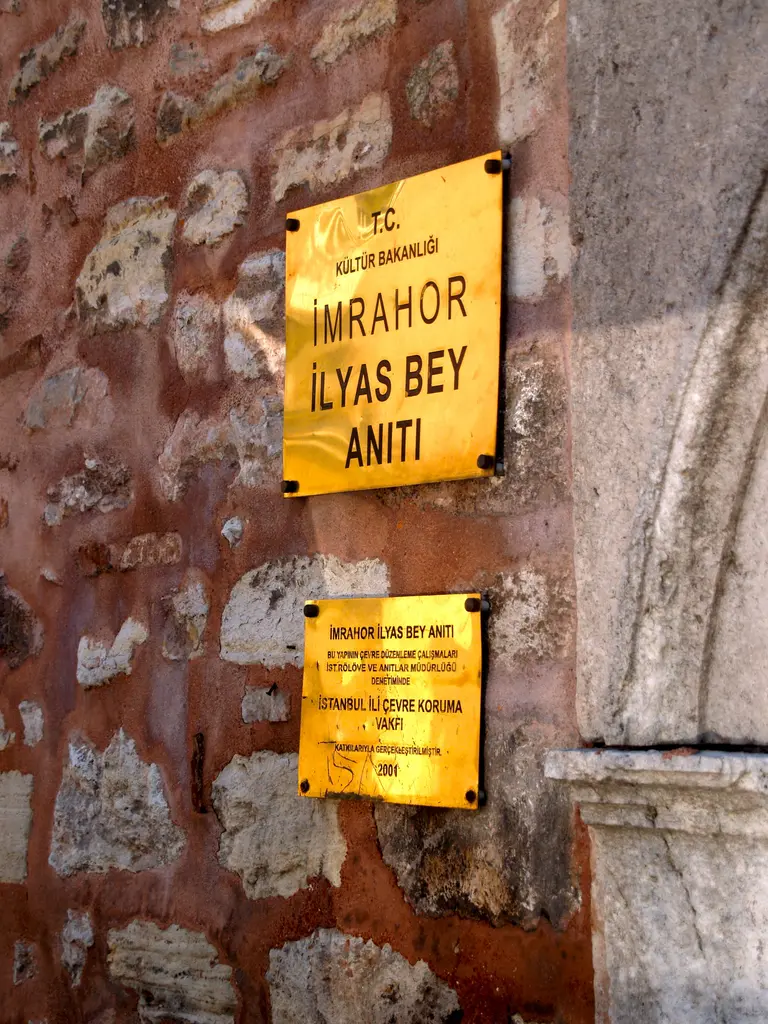 İmrahor İlyas Bey Camii'nin duvarındaki sarı tanıtım levhası; üstte "İmrahor İlyas Bey Anıtı" yazısı, altında küçük bilgi levhası, arkada sıvası dökülmüş tuğla duvar.
