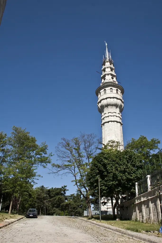 Kampüs bahçesinden Beyazıt Kulesi; ağaçlı yol, kenardaki alçak duvar, arkada tam boy görünen kule ve açık gökyüzü.