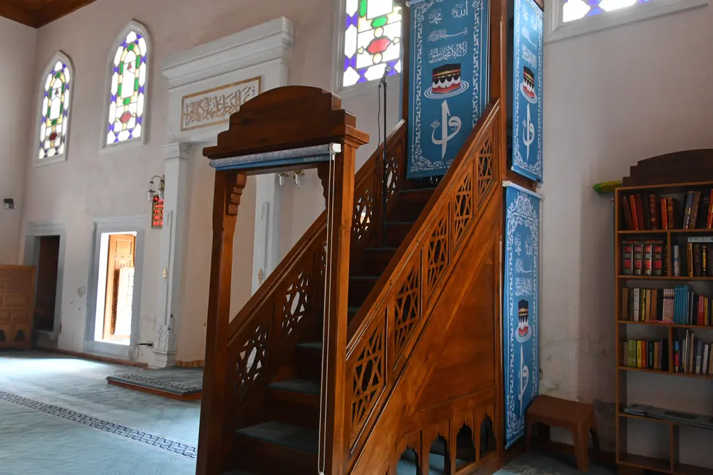 Şah Sultan Camii'nde minber; ahşap merdiven korkulukları, mavi süslemeli örtü, kitaplık ve renkli camlı pencereler.