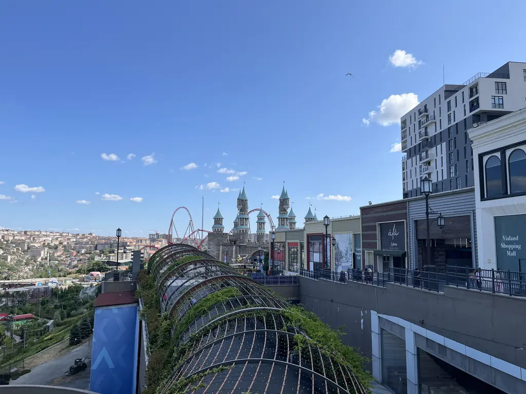 Vialand'de alışveriş merkezinin terasından tema park; yeşil sarmaşıklı kemerli yaya köprüsü, uzakta şato kuleleri ve mahalleler.