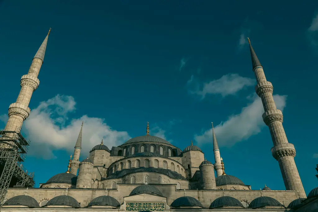 Sultanahmet'te aşağıdan Sultanahmet Camii; koyu mavi gökyüzüne uzanan iki ince minare, kademelenen kurşuni kubbeler ve kemerli pencereli taş cephe.