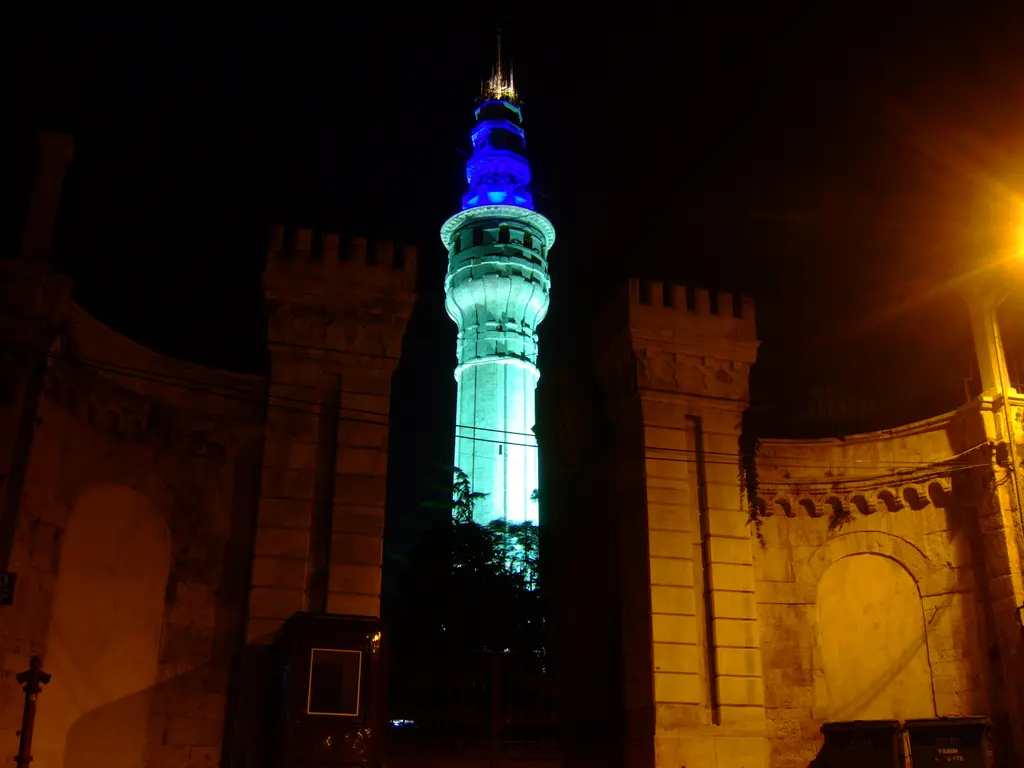 Gece aydınlatmalı Beyazıt Kulesi; mavi ile turkuaza boyanmış gövde, önündeki dişli kapı yapısının turuncu ışığı ve karanlık gökyüzü.